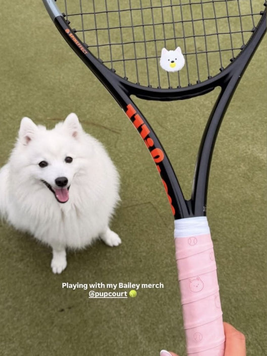 A customer photo. A white pup on a grass court beside a racket fitted with a Pupcourt dampener and a pink Pupcourt overgrip.