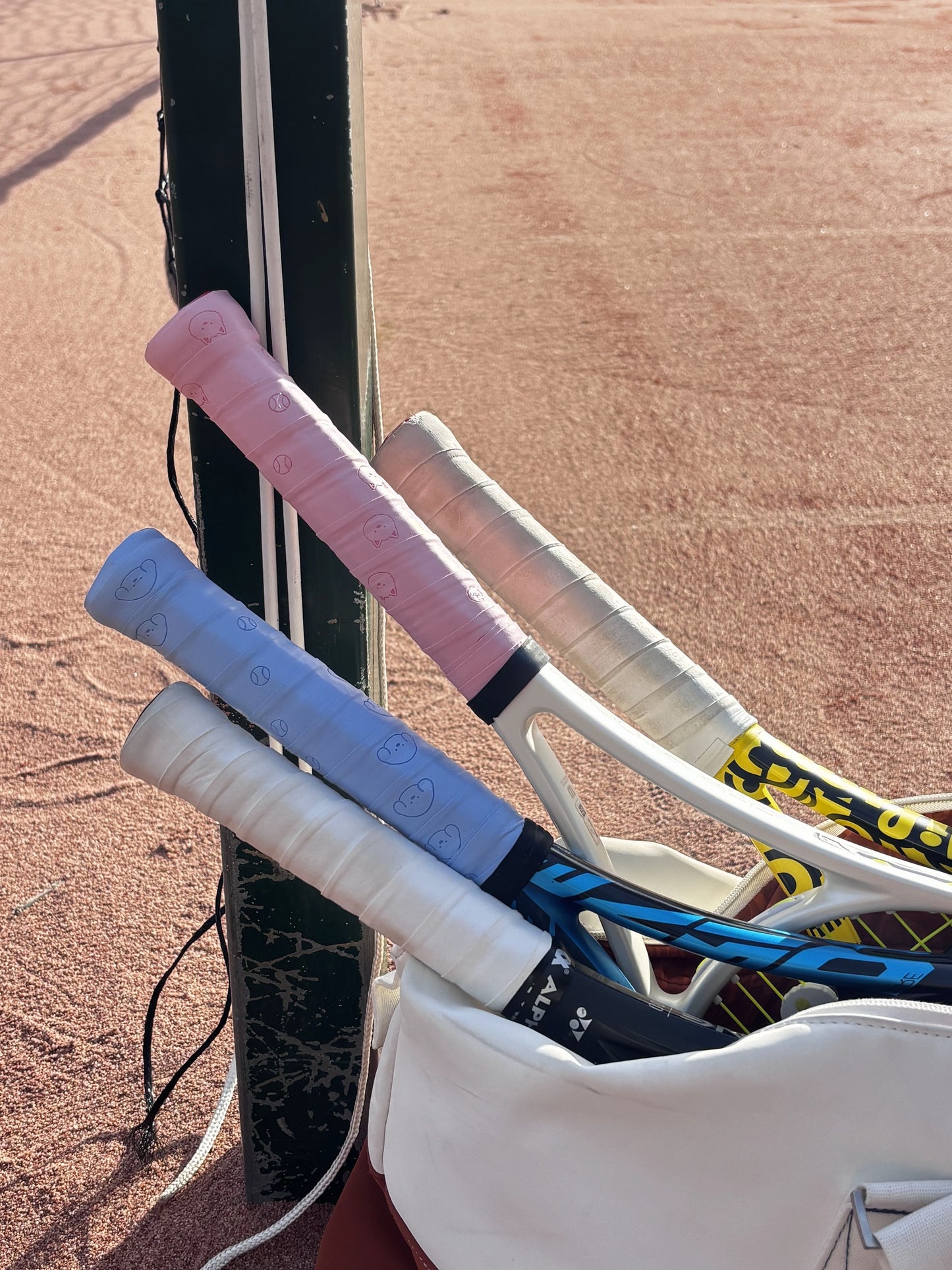 pwp-overgrip-court-2026-08-02 Pupcourt overgrips wrapped on rackets resting against the net post on a clay court, the blue golden retriever print and the pink cavoodle print side by side